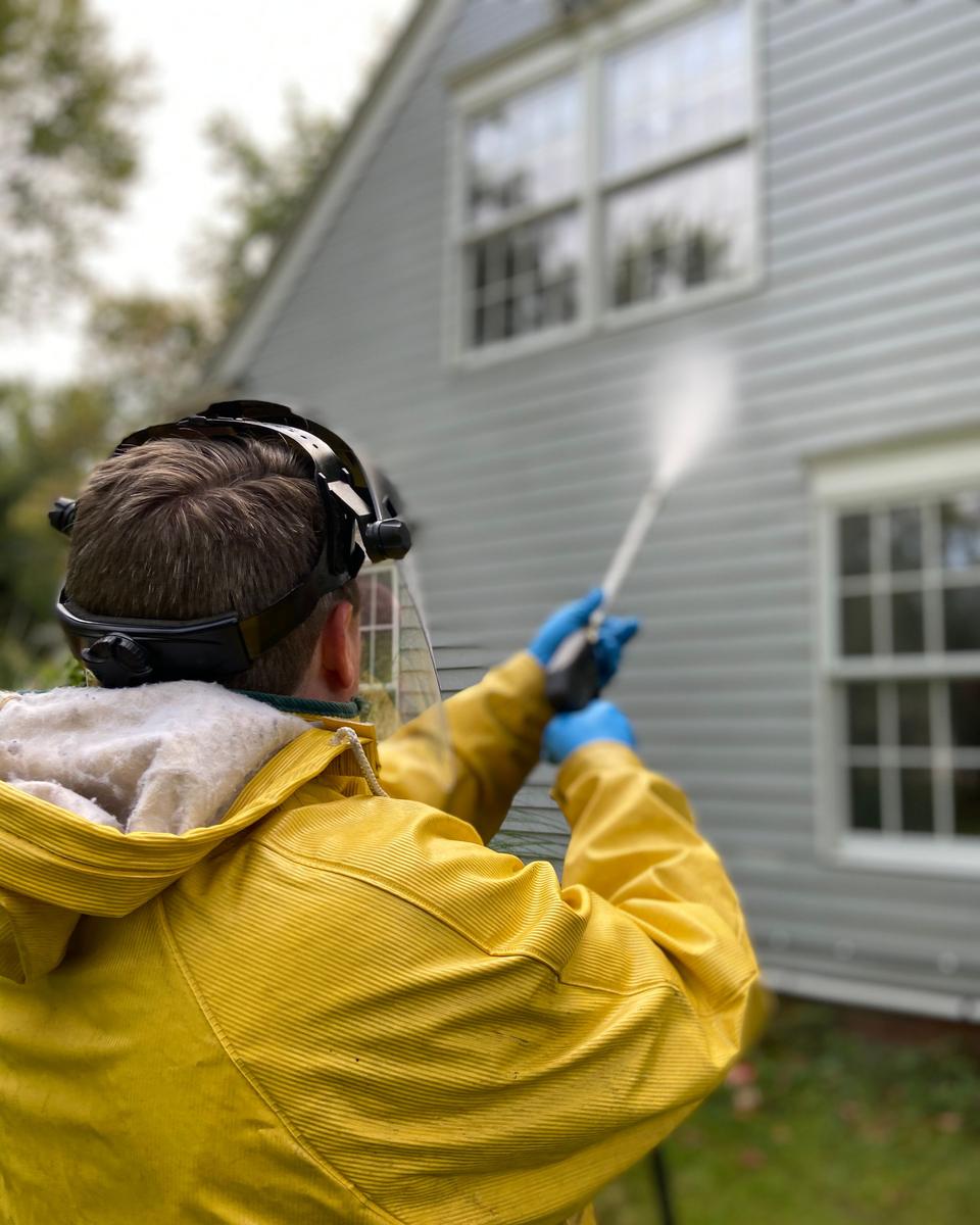 A person in a yellow raincoat pressure washing the siding of a house, seen from behind (representative stock photo)